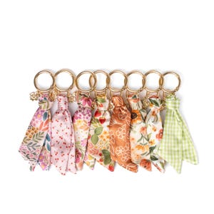 May include: A collection of eight keychains with gold-colored rings and floral patterned fabric tassels. The tassels feature various designs, including flowers, cherries, and gingham, in shades of pink, orange, and green.