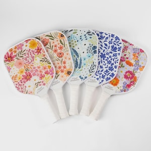 May include: Five pickleball paddles with floral designs in various colors. Each paddle has a white handle and a different floral pattern, including pink, orange, blue, and purple flowers. The brand name "elyse bredanne" is printed on each paddle.