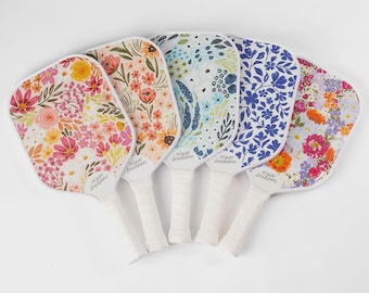 May include: Five pickleball paddles with floral designs in various colors. Each paddle has a white handle and a different floral pattern, including pink, orange, blue, and purple flowers. The brand name "elyse bredanne" is printed on each paddle.