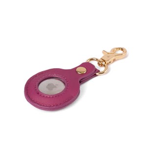 May include: A magenta faux leather protective case for an Apple AirTag featuring a textured surface, a gold-toned metal snap button closure, and a matching gold-toned lobster claw clasp attached to a circular metal ring for secure fastening.