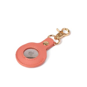May include: A coral-colored textured protective holder for a circular tracking device, featuring a gold-toned metal lobster clasp and a matching gold-toned rivet. The holder has a circular cutout that reveals the metallic surface of the tracker.