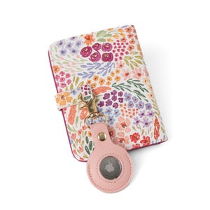 May include: A rectangular wallet featuring a white background with a colorful floral pattern in shades of pink, orange, purple, and green. A light pink circular tracker holder is attached to the wallet with a gold-toned metal lobster clasp.