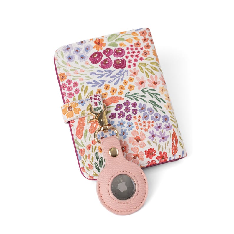 May include: A rectangular wallet featuring a white background with a colorful floral pattern in shades of pink, orange, purple, and green. A light pink circular tracker holder is attached to the wallet with a gold-toned metal lobster clasp.
