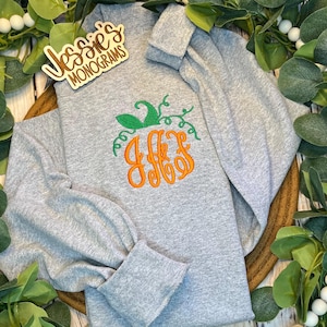 May include: Gray long-sleeve shirt with a pumpkin-shaped monogram featuring the letters "JFF" in orange with green vines. The shirt has a cuffed sleeve and is displayed on a wooden surface with green foliage.