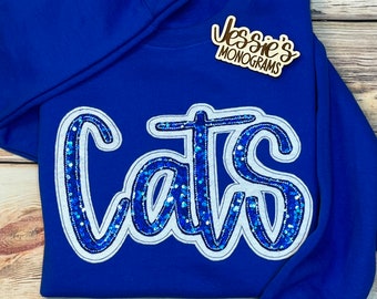 May include: Royal blue sweatshirt with a white outline and blue sequin lettering that reads "Cats". A small wooden sign with the text "Jessie's Monograms" is in the top right corner of the image.