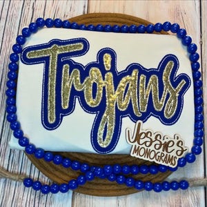 May include: White t-shirt with a blue and gold "Trojans" design. The shirt is decorated with blue beads. A small wooden tag with the text "Jessie's Monograms" is attached to the shirt.
