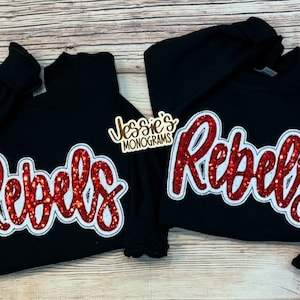 May include: Two black sweatshirts with red glitter "Rebels" lettering. The lettering is outlined in white.