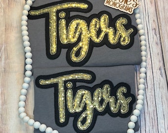 May include: Two grey t-shirts with the word "Tigers" in gold glitter letters on each shirt. The word "Tigers" is outlined in black.