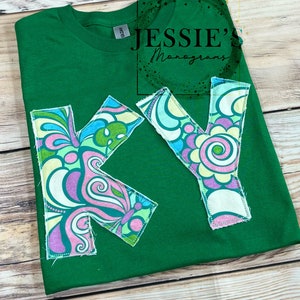 May include: Green t-shirt with a large "KY" design made from colorful fabric patches. The patches feature a swirl pattern and a butterfly design. The shirt has a tag that says "NWTG" and a small logo that says "Jessie's Monograms".