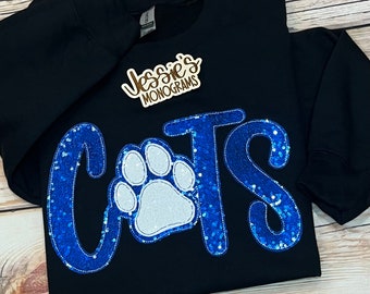 Könnte beinhalten: Schwarzer Pullover mit blauer und weißer Pailletten-Beschriftung "CATS" und einem weißen Pfotenabdruck in der Mitte.