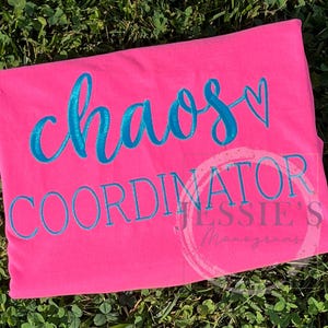 May include: A bright pink t-shirt with the words "chaos coordinator" in turquoise script. A small turquoise heart is next to the word "chaos". The shirt is laying on a bed of green grass.