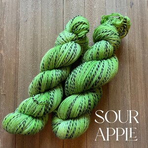 May include: Two skeins of green and black yarn with a speckled pattern. The yarn is labeled "Sour Apple".