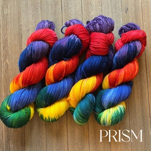 May include: Four skeins of yarn in a rainbow color gradient. The yarn is a mix of red, orange, yellow, green, blue, and purple. The yarn is arranged in a criss-cross pattern. The yarn is labeled "PRISM".
