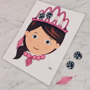 Puede incluir: Una actividad de manualidades imprimible con la cara de una niña con una corona rosa, un collar y pendientes. La actividad incluye un espacio para un nombre y tres pegatinas de gemas blancas y negras.