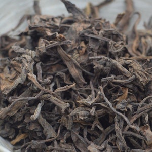 以下が含まれることがあります： 小さな灰色の陶器のボウルに入った、茶色の茶葉の束のクローズアップ。