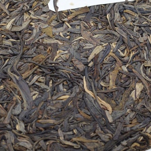 May include: A round, compressed tea cake with a dark brown color, composed of tightly packed tea leaves. A white label with black text and a drawing is placed on top of the tea cake. The tea cake is wrapped in white paper.