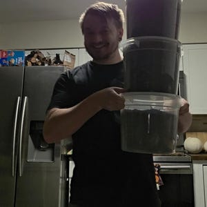 May include: A person holding three clear plastic containers stacked on top of each other. The containers are filled with a dark substance. The person is wearing a black t-shirt and smiling. The background includes a refrigerator and kitchen cabinets.