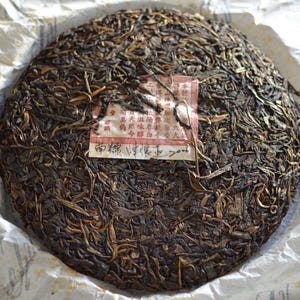 May include: A round, compressed tea cake with a dark brown color, wrapped in white paper. The tea cake has a textured surface with visible tea leaves. A small, rectangular label with Chinese characters is attached to the tea cake.