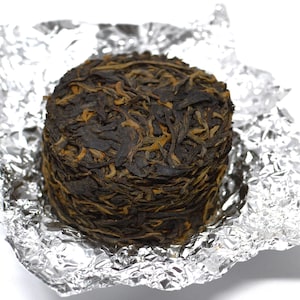 Puede incluir: Un pastel de té redondo y comprimido, de color marrón oscuro con reflejos dorados, se presenta sobre un envoltorio de papel de aluminio plateado arrugado. El pastel de té tiene un aspecto texturizado y en capas, lo que sugiere hojas de té muy compactas.