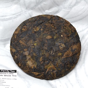 May include: A circular compressed cake of dark brown and green tea leaves. A white label reads: Wang Family Tea, Sun Moon Lake, Red Jade White Tea, Net Weight: 200Grams ± 15g, MFD: 2026.04.22, MADE IN TAIWAN, BATCH number wildgarden2026SPRING.