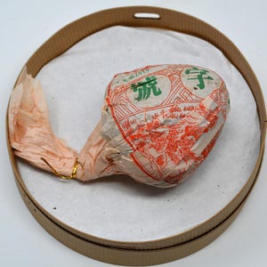 May include: A round, brown paper-wrapped tea cake with Chinese characters and a red and white design. The cake is sitting on a white surface inside a brown cardboard tray.