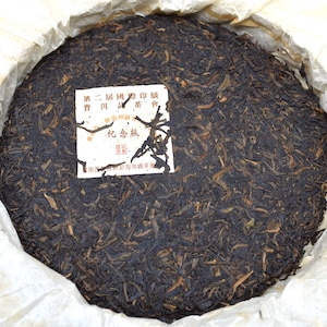 May include: A round, compressed tea cake with a dark brown color, wrapped in off-white paper. A white label with Chinese characters is affixed to the tea cake. The tea cake is a traditional Chinese tea product.