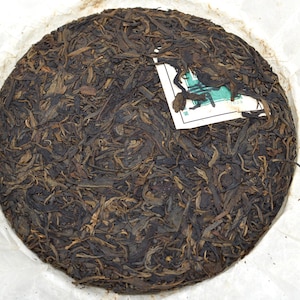 May include: A round, compressed tea cake with a dark brown color, wrapped in white paper. The tea leaves are tightly packed and have a textured appearance. A small label is visible on the surface.
