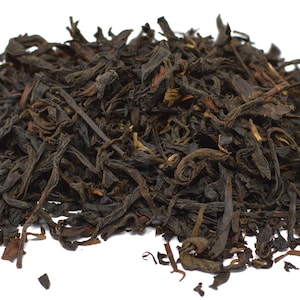 May include: A close-up shot of a pile of loose-leaf tea. The tea leaves are dark brown and black, with a slightly curled and twisted appearance. The tea is set against a plain white background.