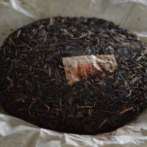 May include: A round, compressed tea cake, dark brown in color, wrapped in off-white paper. The tea cake has a textured surface with visible tea leaves and stems. A small, rectangular label is affixed to the cake.