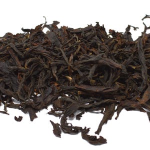 May include: A pile of loose-leaf black tea consisting of dark brown, curled, and twisted dried tea leaves. The texture is brittle and irregular, with varying shades of deep mahogany and near-black tones throughout the scattered tea leaf collection.