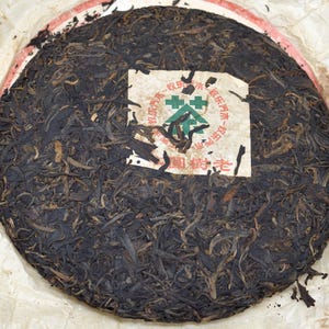 May include: A round, dark brown tea cake with a paper label. The tea cake is wrapped in paper. The label has Chinese characters and a green and white design. The tea leaves are tightly compressed.