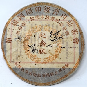 May include: A round, aged tea cake with Chinese characters and ornate designs. The tea cake has a faded appearance with a central design in orange and brown, surrounded by text in red and black. The outer edge is a darker gray.