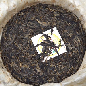 Puede incluir: Un pastel redondo y comprimido de hojas de té de color marrón oscuro, envuelto en papel blanquecino. Una pequeña etiqueta blanca con detalles amarillos y rojos es visible en la parte superior. Las hojas de té tienen una apariencia texturizada.