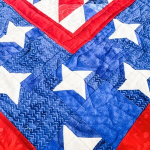 Op de afbeelding: Close-up van een gewatteerd textiel met een patriottisch ontwerp. De quilt toont een levendig kleurenpalet van rood, wit en blauw. Witte sterren zijn geplaatst tegen een blauwe achtergrond, met rode randen en accenten. De stiksels voegen textuur toe.