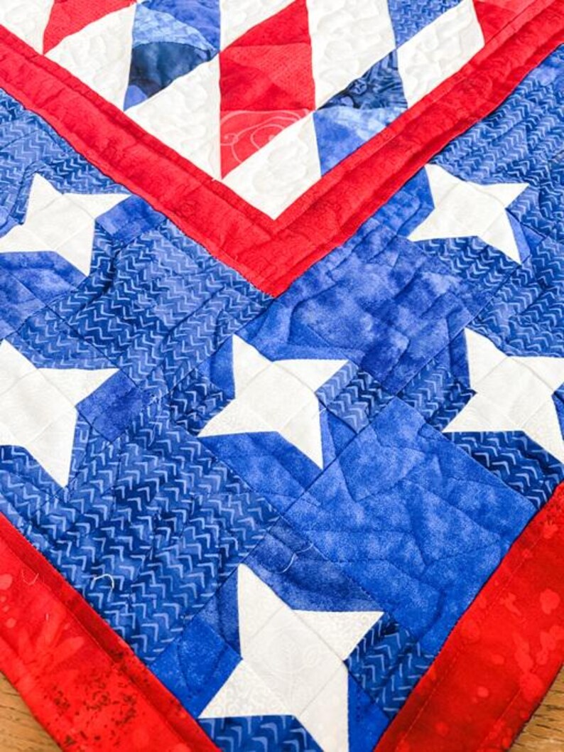 Op de afbeelding: Close-up van een gewatteerd textiel met een patriottisch ontwerp. De quilt toont een levendig kleurenpalet van rood, wit en blauw. Witte sterren zijn geplaatst tegen een blauwe achtergrond, met rode randen en accenten. De stiksels voegen textuur toe.