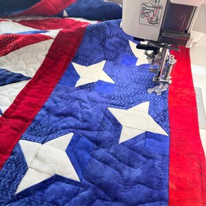 Op de afbeelding: Een close-up van een quilt in wording, met een blauw veld met witte sterren en een rode rand. De quilt wordt genaaid met een witte naaimachine. De stof heeft een gewatteerd patroon.