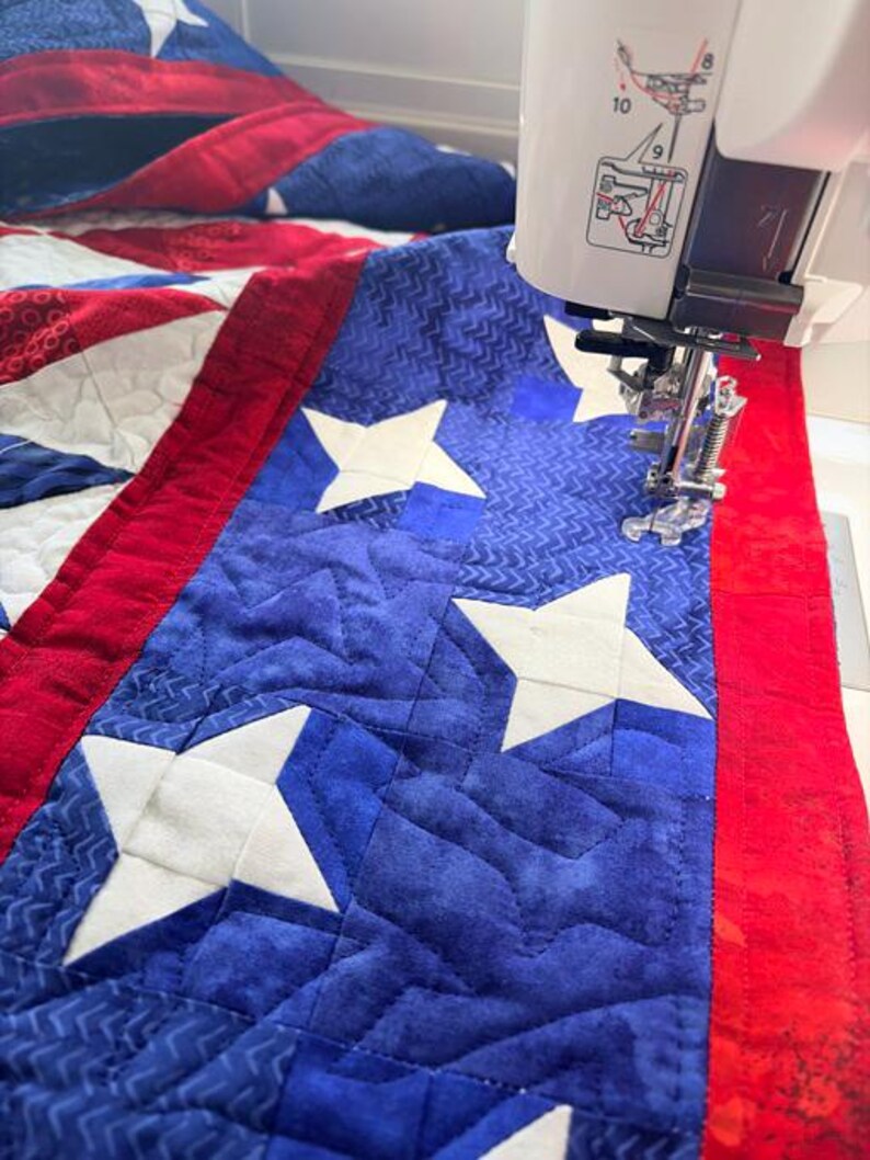 Op de afbeelding: Een close-up van een quilt in wording, met een blauw veld met witte sterren en een rode rand. De quilt wordt genaaid met een witte naaimachine. De stof heeft een gewatteerd patroon.