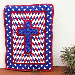 Op de afbeelding: Een patriottische quilt met een groot blauw kruis in het midden, omgeven door een patroon van rode, witte en blauwe ruitvormen. De rand is blauw met witte sterren en een rode rand. De quilt hangt tegen een witte muur.