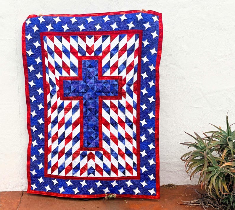 Op de afbeelding: Een patriottische quilt met een groot blauw kruis in het midden, omgeven door een patroon van rode, witte en blauwe ruitvormen. De rand is blauw met witte sterren en een rode rand. De quilt hangt tegen een witte muur.