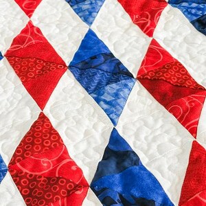 Op de afbeelding: Close-up van een quilt met een ruitpatroon in rode en blauwe stoffen. De ruiten zijn geplaatst tegen een witte achtergrond met gewatteerde stiksels. De rode ruiten hebben een wervelpatroon, terwijl de blauwe een patroonontwerp hebben.