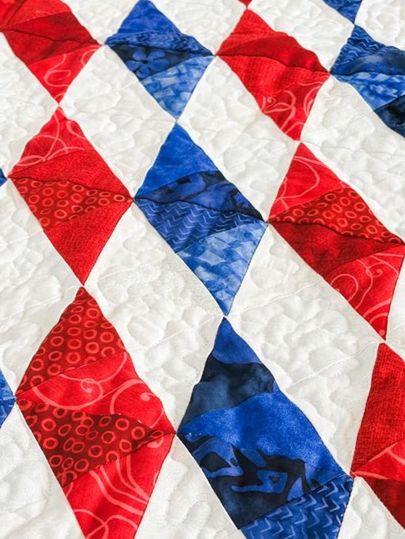 Op de afbeelding: Close-up van een quilt met een ruitpatroon in rode en blauwe stoffen. De ruiten zijn geplaatst tegen een witte achtergrond met gewatteerde stiksels. De rode ruiten hebben een wervelpatroon, terwijl de blauwe een patroonontwerp hebben.