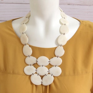 May include: A cream-coloured statement necklace featuring large, oval-shaped beads connected by black string. The necklace is displayed on a mannequin, set against a mustard-yellow blouse and a light wood background. The necklace has a layered design.