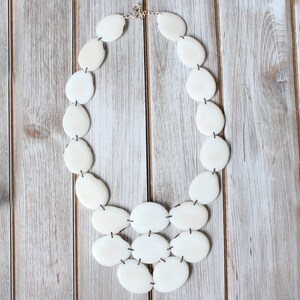 May include: Cream-coloured statement necklace with irregularly shaped, oval beads. The beads are strung together with dark string and connected with a silver clasp. The necklace is displayed on a light-coloured wooden surface.