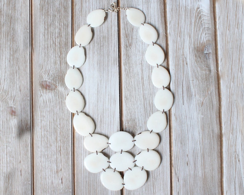 May include: Cream-coloured statement necklace with irregularly shaped, oval beads. The beads are strung together with dark string and connected with a silver clasp. The necklace is displayed on a light-coloured wooden surface.
