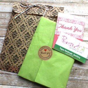 May include: Overhead view of a parcel wrapped in green tissue paper, a brown damask patterned bag, and two cards. One card reads "Thank You" in pink script, and the other reads "PERFECTOH" with "Tagua Handmade Jewellery" below it.