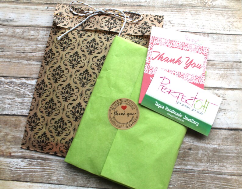 May include: Overhead view of a parcel wrapped in green tissue paper, a brown damask patterned bag, and two cards. One card reads "Thank You" in pink script, and the other reads "PERFECTOH" with "Tagua Handmade Jewellery" below it.
