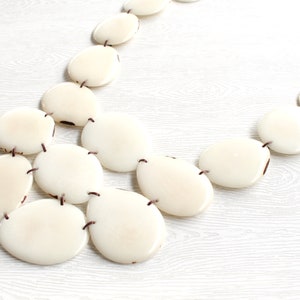 May include: Cream-coloured statement necklace with large, oval beads. The beads are linked by dark brown string, forming a layered design. The necklace is shown on a light-coloured wooden surface, accentuating the creamy tones of the beads.