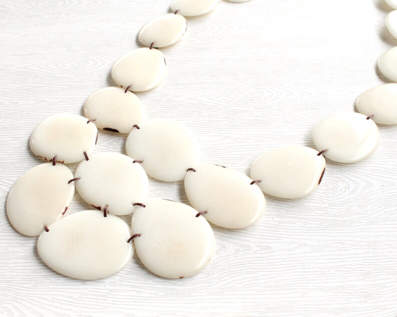 May include: Cream-coloured statement necklace with large, oval beads. The beads are linked by dark brown string, forming a layered design. The necklace is shown on a light-coloured wooden surface, accentuating the creamy tones of the beads.