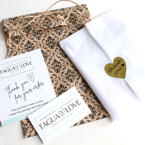 May include: A white paper bag with a brown patterned design. The bag is tied with twine and has a small gold heart-shaped sticker with the text "thank you". The bag is sitting on a white surface. The bag is also decorated with two white cards with the text "TAGUA LOVE" and "Thank you for your order".