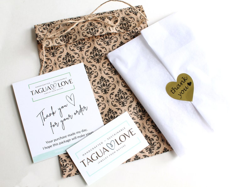 May include: A white paper bag with a brown patterned design. The bag is tied with twine and has a small gold heart-shaped sticker with the text "thank you". The bag is sitting on a white surface. The bag is also decorated with two white cards with the text "TAGUA LOVE" and "Thank you for your order".